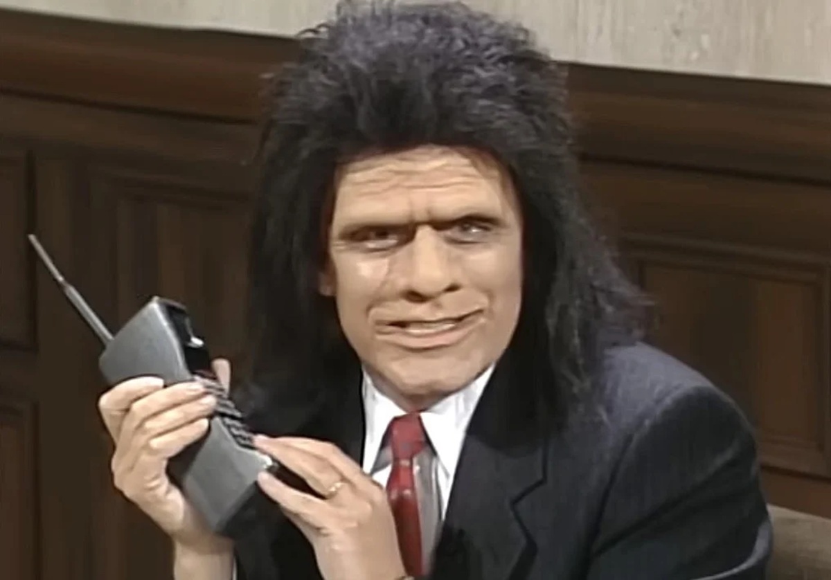 Man in a Suit with a Long Black Wig Holding a Large Brick style Mobile Phone with an Antenna in a Wood paneled Room