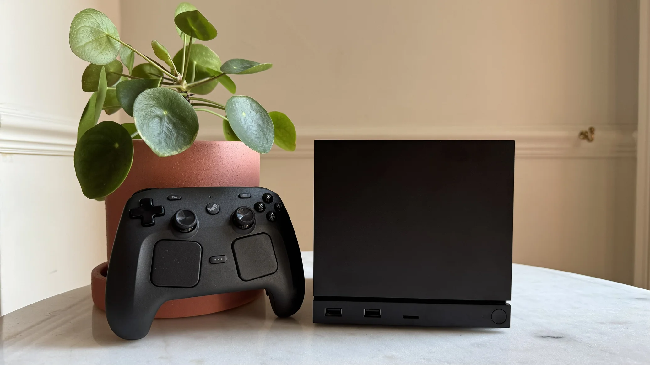 Black Gaming Console with a Wireless Controller on a White Tabletop Beside a Terracotta Pot with a Leafy Plant